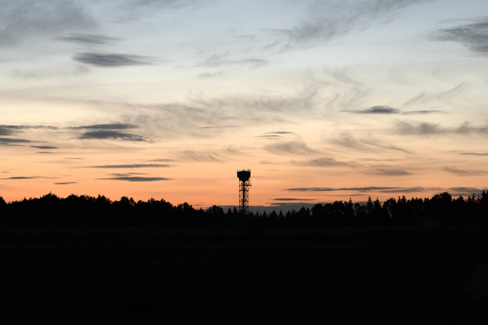 The image depicts a serene sunset over a field, with a tall tower standing in the center and silhouetted against the warm hues of orange and pink sky. The sun is setting behind the trees on either side of the tower, casting long shadows across the landscape. The scene conveys a sense of tranquility and peacefulness as the day comes to an end.