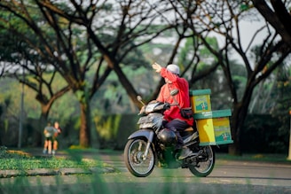 A person wearing a bright red jacket and a hat is riding a motorcycle with a delivery box on the back. The individual is extending their left hand, possibly to signal or wave. In the background, two people are jogging along a path, surrounded by tall trees with lush green foliage.
