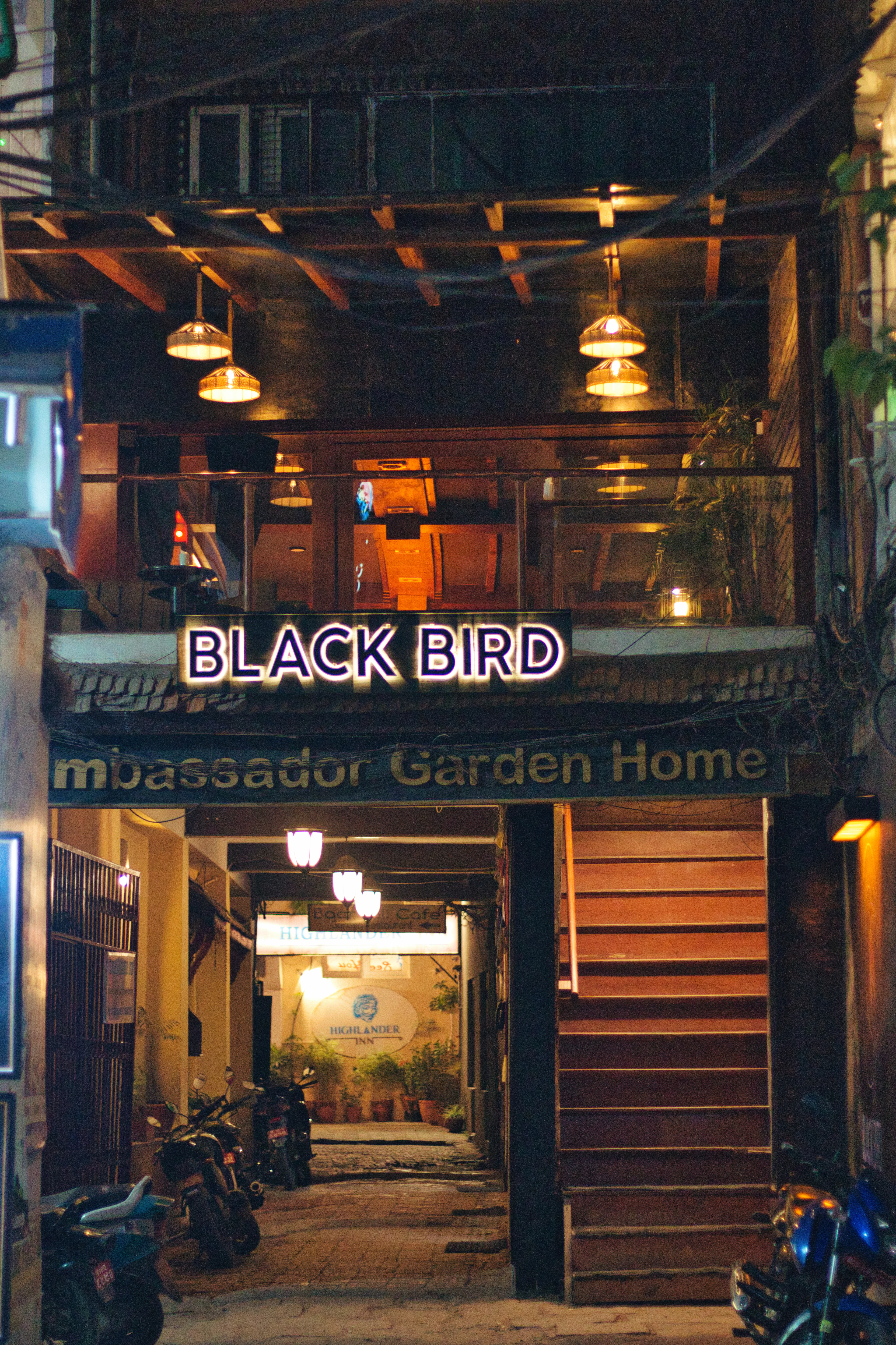 A warmly lit first-floor cafe terrace in Kathmandu at night, its neon sign glowing above a shadowed lane of parked motorbikes and a lamplit courtyard passage behind
