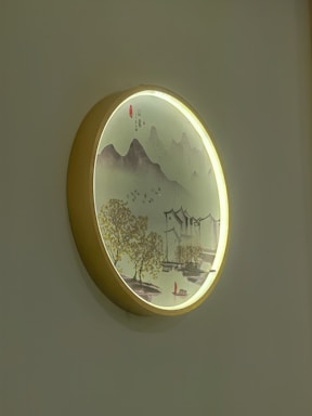 A round wall-mounted art piece featuring an elegant landscape scene. It depicts mountains, trees, a traditional building, and a boat, all highlighted by soft lighting from the frame.