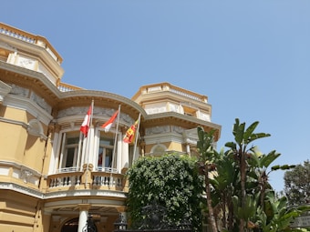 A grandiose, historical building with intricate architectural details featuring columns, ornate moldings, and decorative elements. The facade is painted in a warm, pastel yellow with white trim. Three flags are mounted on poles above the entrance, possibly indicating national or organizational affiliation. Lush green foliage, including palm trees and other tropical plants, adorns the surroundings under a clear blue sky.
