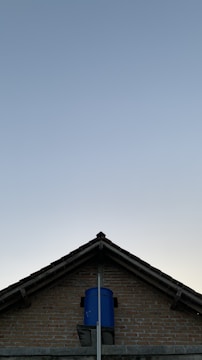 A triangular brick house roof with a blue water tank installed at its top-center against a clear sky backdrop.