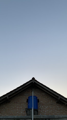 A triangular brick house roof with a blue water tank installed at its top-center against a clear sky backdrop.