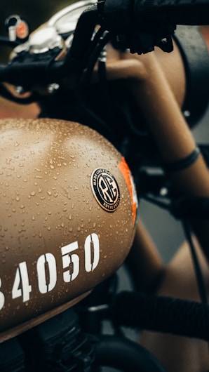 A close-up view of a motorcycle fuel tank, covered in water droplets, featuring a beige color with a distinct logo and the number '34050' in white. The handlebars and other parts of the motorcycle are visible, showing a classic design.