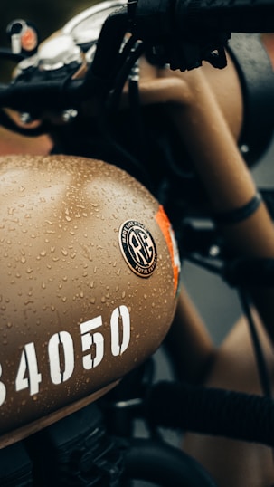 A close-up view of a motorcycle fuel tank, covered in water droplets, featuring a beige color with a distinct logo and the number '34050' in white. The handlebars and other parts of the motorcycle are visible, showing a classic design.