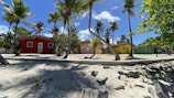 Colorful houses with thatched roofs sit amidst tall palm trees on a sandy beach. The sky is clear with a few scattered clouds, and shadows of the trees stretch across the sand.