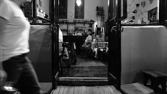 An indoor setting of a rustic cafe or restaurant with wooden floors and dim lighting. People are seated at tables in the background while a person in motion is photographed in the foreground, adding a dynamic element to the scene. The walls are decorated with shelves and various wall hangings.