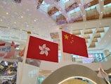Two flags are prominently displayed indoors against a backdrop of a modern shopping mall or public area. The flags are the Hong Kong flag on the left and the China flag on the right. The ceiling features intricate, decorative lighting resembling snowflakes, and the area is spacious with wooden accents on the ceilings.