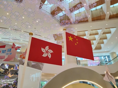 Two flags are prominently displayed indoors against a backdrop of a modern shopping mall or public area. The flags are the Hong Kong flag on the left and the China flag on the right. The ceiling features intricate, decorative lighting resembling snowflakes, and the area is spacious with wooden accents on the ceilings.
