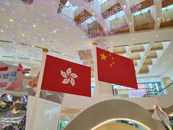Two flags are prominently displayed indoors against a backdrop of a modern shopping mall or public area. The flags are the Hong Kong flag on the left and the China flag on the right. The ceiling features intricate, decorative lighting resembling snowflakes, and the area is spacious with wooden accents on the ceilings.