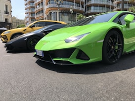 Three luxury sports cars are parked on a street with modern architecture in the background. The cars are different colors: bright green, black, and yellow. The setting appears urban with a mix of concrete surfaces and greenery.