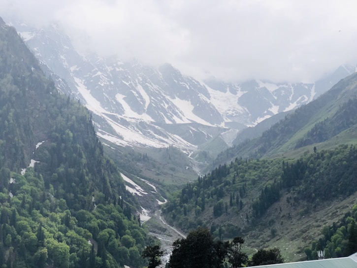 Snow-capped mountains tower in the background, surrounded by misty clouds. A lush green valley with dense forested slopes stretches out below, with a narrow winding river cutting through the landscape.