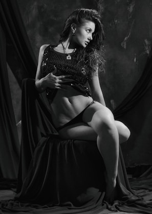 A black and white portrait of a woman in a seated pose, wearing a dark, sleeveless, sparkling top and black underwear. She has long, flowing hair and is looking to the side with a relaxed, confident expression. The backdrop features dark drapery, enhancing the dramatic lighting and shadows.