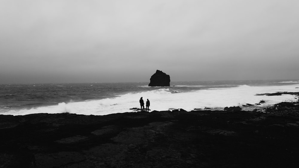 a couple of people standing on a beach by a large rock