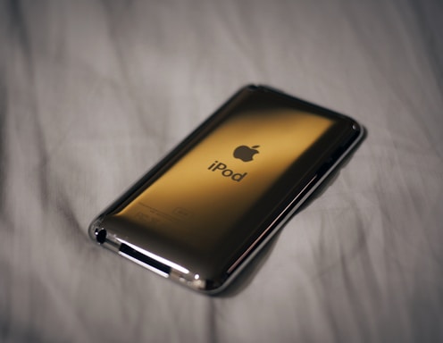 A back view of an iPod with an Apple logo and the word 'iPod' prominently displayed in the center. It is resting on a soft, white fabric surface, and the lighting casts a smooth reflection on the metallic body of the device.