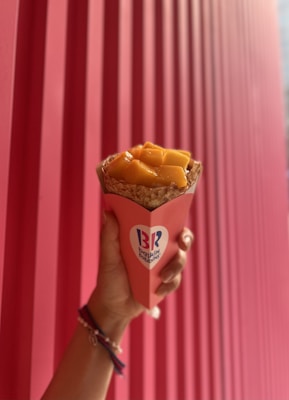 A hand is holding a crepe filled with orange fruit cubes, wrapped in a pink and red Baskin Robbins branded paper against a pink corrugated background.