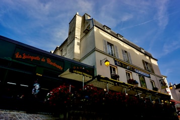 A multi-story building with charming architectural design stands against a clear blue sky. The building includes businesses, with awnings and signage in French. One establishment, La Guinguette de Montmartre, has a nostalgic feel with posters and outdoor seating. Another establishment, La Bonne Franquette, features outdoor seating, street lamps, and flower arrangements.