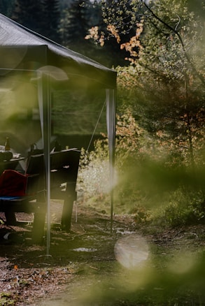 A tranquil outdoor setting featuring a canopy tent partially visible among lush greenery. Dappled sunlight filters through the leaves, creating a serene atmosphere. A picnic table and benches are under the tent, with some items like a bottle and bag placed on them. The scene suggests a peaceful retreat into nature, surrounded by trees and undergrowth.