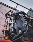 A close-up view of a collection of miscellaneous items bundled together and mounted on a wall. The objects include a remote control, a power strip, various cables, and other electronic devices, all secured with a strap. The scene is outdoors with a cloudy sky visible in the background.