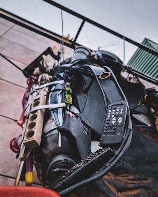 A close-up view of a collection of miscellaneous items bundled together and mounted on a wall. The objects include a remote control, a power strip, various cables, and other electronic devices, all secured with a strap. The scene is outdoors with a cloudy sky visible in the background.