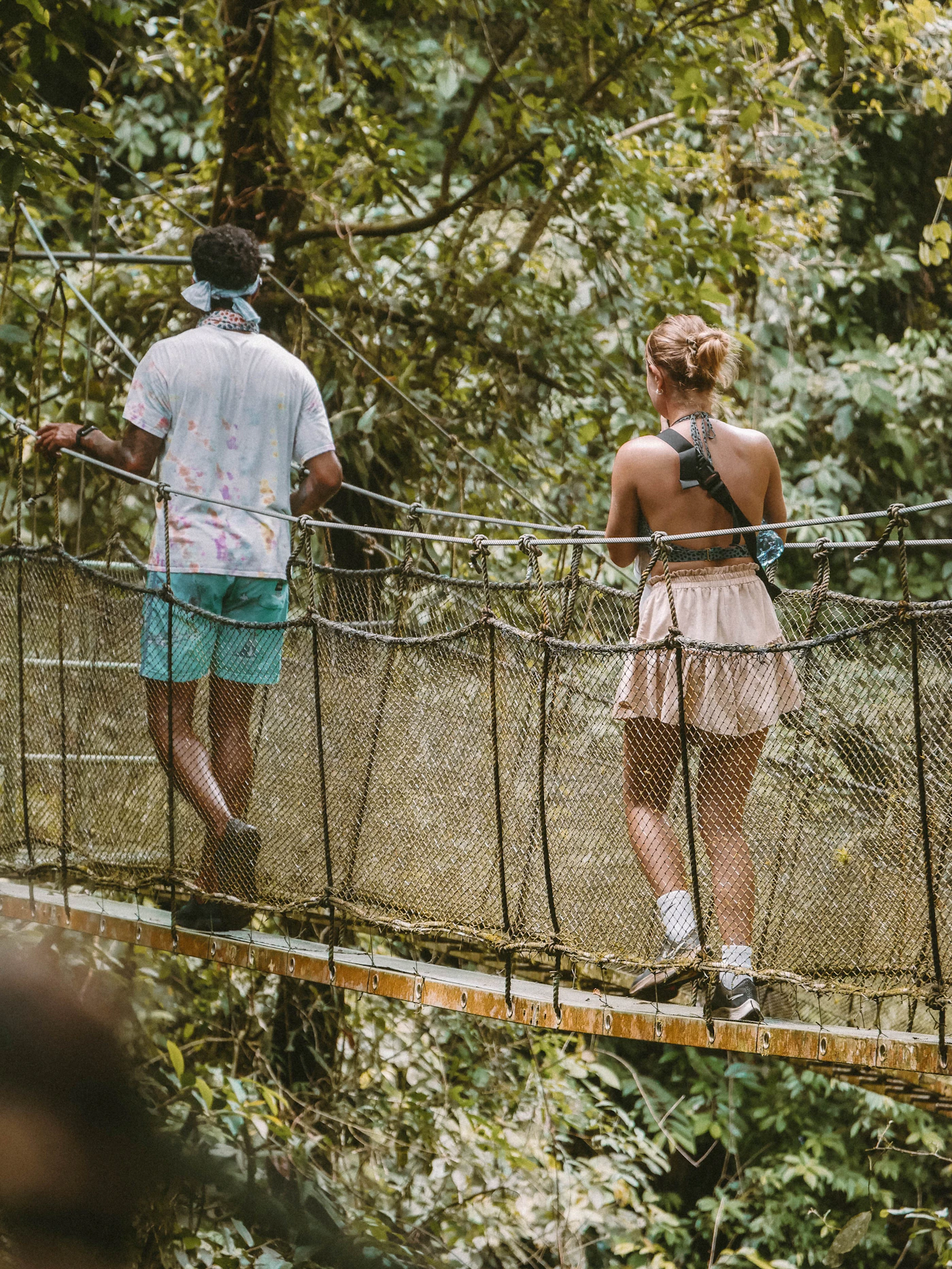 A couple walking across a wooden suspension bridge in Costa Rica high above the jungle canopy with green forest stretching in every direction below and mountains visible in the background