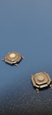 Two small metallic tactile switches are positioned on a textured blue surface. Each switch has a white button in the center and several protruding legs for mounting.