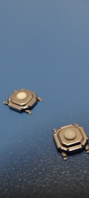 Two small metallic tactile switches are positioned on a textured blue surface. Each switch has a white button in the center and several protruding legs for mounting.