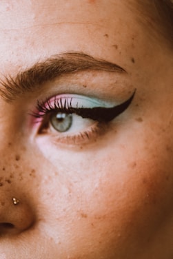 A close-up of the left eye area with detailed makeup featuring a blend of pastel blue and pink eyeshadow, accentuated by a bold black winged eyeliner. The skin is fair with visible freckles, and there is a small nose piercing on the side.