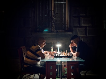 Two elderly men are deeply engaged in a game of chess, sitting across from each other at a small glass table. The scene is dimly lit by a single overhead lamp, creating a focused and intimate setting. A bottle, possibly of water or a beverage, is placed on the table beside the chessboard, and the background shows a dark, textured wall with a window.