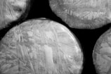 Several round objects wrapped in reflective plastic material, with visible crisscross patterns and varying textures. The image is in black and white, highlighting contrast and detail in the wrapping.