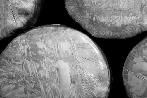 Several round objects wrapped in reflective plastic material, with visible crisscross patterns and varying textures. The image is in black and white, highlighting contrast and detail in the wrapping.