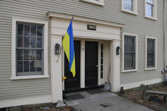 A two-story building with beige siding and multiple windows. A plaque with the number 1803 is above the black double doors. A blue and yellow flag is mounted beside the entrance. A snow shovel rests against the wall next to the door.