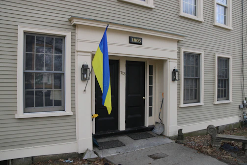 A two-story building with beige siding and multiple windows. A plaque with the number 1803 is above the black double doors. A blue and yellow flag is mounted beside the entrance. A snow shovel rests against the wall next to the door.