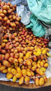 A large pile of ripe dates with vibrant yellow and orange hues is displayed on a bed of newspaper. Some green plastic bags are partially visible in the top right corner. The dates are in clusters, still attached to their stems.