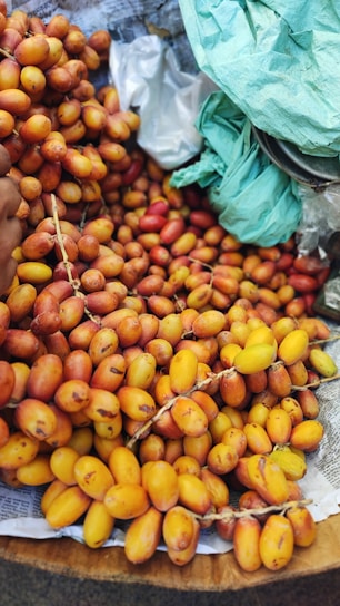 A large pile of ripe dates with vibrant yellow and orange hues is displayed on a bed of newspaper. Some green plastic bags are partially visible in the top right corner. The dates are in clusters, still attached to their stems.