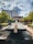A tranquil outdoor scene features a long water fountain bordered by greenery. In the background, a domed structure is surrounded by modern office buildings. The sky is partly cloudy with patches of sunlight illuminating the landscape.