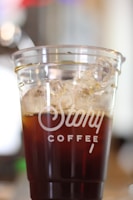A close-up of a transparent cup filled with iced coffee, branded with Cafecup logo