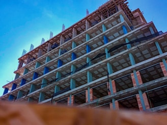 A tall, unfinished concrete building structure with multiple floors is shown from a low angle against a clear blue sky. The building's framework includes visible pillars, open spaces for windows, and construction materials. Some scaffolding is apparent on the roof, indicating ongoing construction work.