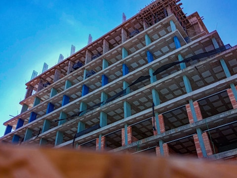 A tall, unfinished concrete building structure with multiple floors is shown from a low angle against a clear blue sky. The building's framework includes visible pillars, open spaces for windows, and construction materials. Some scaffolding is apparent on the roof, indicating ongoing construction work.
