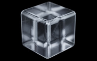 A transparent, glass-like cube with intricate reflections and refractions is placed against a black background. The cube has clean edges and shows subtle internal structures that resemble lines or fibers.