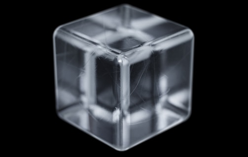 A transparent, glass-like cube with intricate reflections and refractions is placed against a black background. The cube has clean edges and shows subtle internal structures that resemble lines or fibers.