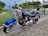 A blue Harley-Davidson motorcycle is parked on a concrete driveway. The bike features chrome accents and a black leather seat. It is positioned against a backdrop of a suburban neighborhood with a wire fence, green trees, and residential houses in the background.