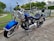 A blue Harley-Davidson motorcycle is parked on a concrete driveway. The bike features chrome accents and a black leather seat. It is positioned against a backdrop of a suburban neighborhood with a wire fence, green trees, and residential houses in the background.