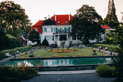 An elegant mansion with red roofs and teal shutters stands behind a formal garden. The garden features symmetrical pathways and a reflecting pool in the foreground, bordered by colorful flower beds and neatly trimmed hedges. Large trees frame the scene, adding lush greenery.