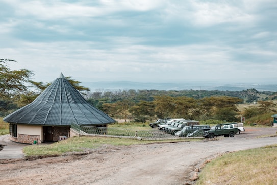A conical-roofed building situated in a grassy landscape with a row of parked safari vehicles nearby. The area is surrounded by lush trees, and the horizon features expansive views of distant hills under a cloudy sky.