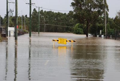 A road submerged under floodwater with a warning sign indicating 'Water Over Road' in the center. Surrounding the road are utility poles and trees, with a line of fencing and greenery extending into the distance.