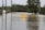 A road submerged under floodwater with a warning sign indicating 'Water Over Road' in the center. Surrounding the road are utility poles and trees, with a line of fencing and greenery extending into the distance.