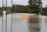 A road submerged under floodwater with a warning sign indicating 'Water Over Road' in the center. Surrounding the road are utility poles and trees, with a line of fencing and greenery extending into the distance.