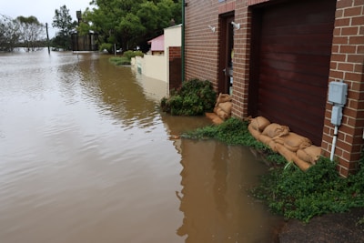 A flooded residential area with water covering the street and reaching up to the garages of brick-faced houses. Sandbags are placed in front of the doors to block water entry. Trees and shrubs are visible drenched in water, and the sky appears overcast, indicating ongoing or recent heavy rainfall.