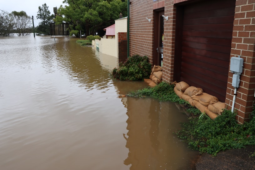 A flooded residential area with water covering the street and reaching up to the garages of brick-faced houses. Sandbags are placed in front of the doors to block water entry. Trees and shrubs are visible drenched in water, and the sky appears overcast, indicating ongoing or recent heavy rainfall.